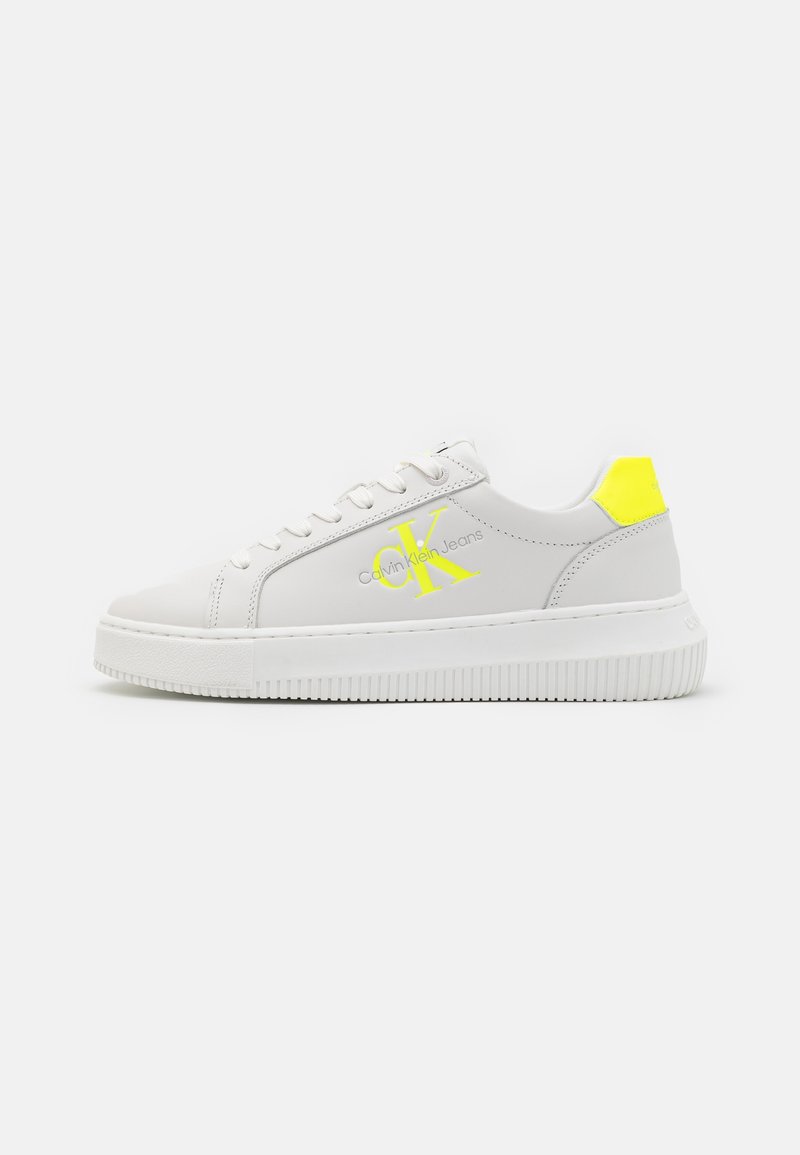 Baskets blanches basses avec semelle épaisse, languette de talon jaune néon et logo jaune "CK Calvin Klein Jeans" sur le côté.