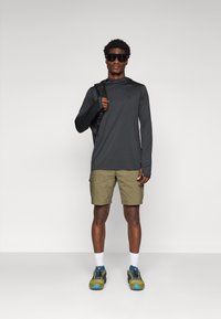 Long-sleeve dark gray shirt, olive green cargo shorts, and multicolor athletic shoes. Accessories include a black backpack and sunglasses.