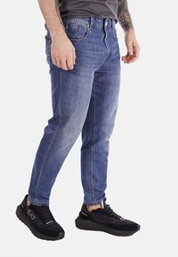 Jeans in denim blu sbiadito con vestibilità tapered, cinque tasche e chiusura con bottoni classica, abbinati a scarpe da ginnastica nere in rete.