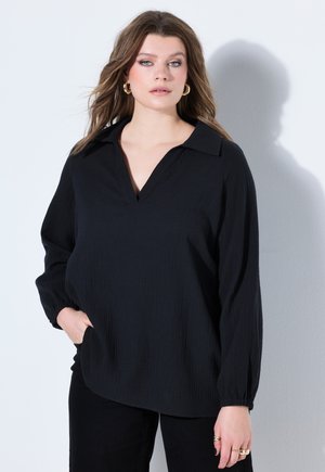 Młoda kobieta o długich brązowych włosach, nosząca złote kolczyki koła, luźną czarną bluzkę z dekoltem w serek i czarne spodnie, stojąca na tle białej ściany.