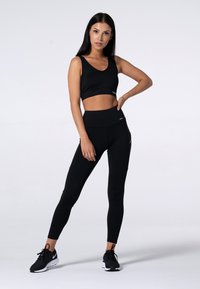 Černý sportovní crop top a legíny s vysokým pasem, vyrobené z pružné látky; modelka pózuje s jednou rukou na boku, na nohou má černé tenisky.
