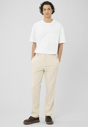 Jeune homme debout les mains dans les poches, portant une chemise blanche à manches courtes, un pantalon beige et des chaussures marron foncé sur un fond uni.