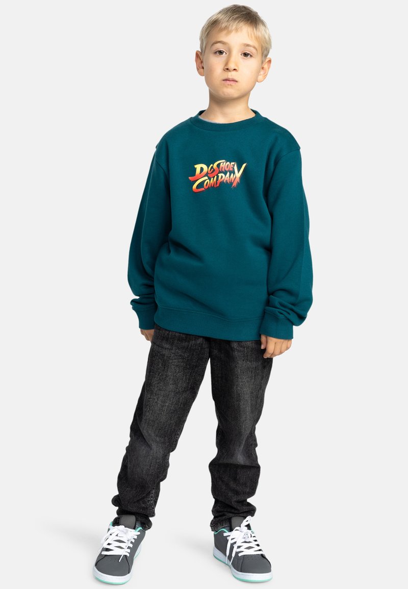 Tealfärgad sweatshirt med "DC Shoe Company"-logotyp i fet stil, i kombination med mörka denimjeans och grå sneakers med vita snören.
