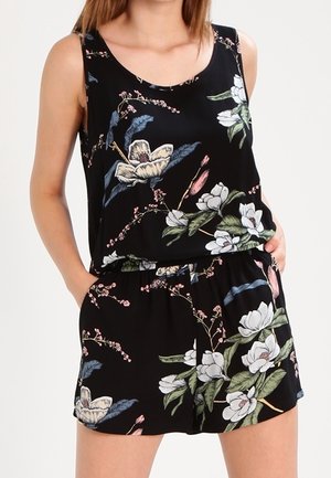 Kvinde iført sort ærmeløs blomstret top og matchende shorts med hvide, grønne og lyserøde blomster, med hænderne i lommerne.