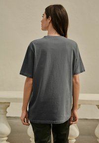 Sivý bavlnený tričko s krátkymi rukávmi, uvoľnený strih, okrúhly výstrih a detaily s surovým okrajom. Viditeľná chrbát modela, v kombinácii s tmavými nohavicami.