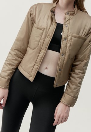 Frau trägt eine beige kurze gesteppte Jacke über einem schwarzen Sport-BH und schwarzen hoch taillierten Leggings, die Hände entspannt an den Seiten.