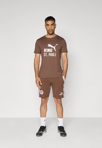 Brun bomulds T-shirt med hvid tekst "KING ST. PAULI", parret med matchende shorts med sidelogoer og accenter. Sorte sportsko og hvide sokker.
