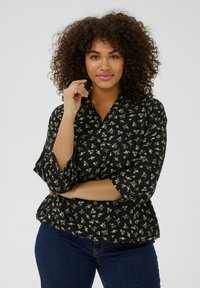Blouse peplum noire avec un motif floral blanc, décolleté en V et manches 3/4. Le tissu a une texture douce, adaptée à un usage décontracté.