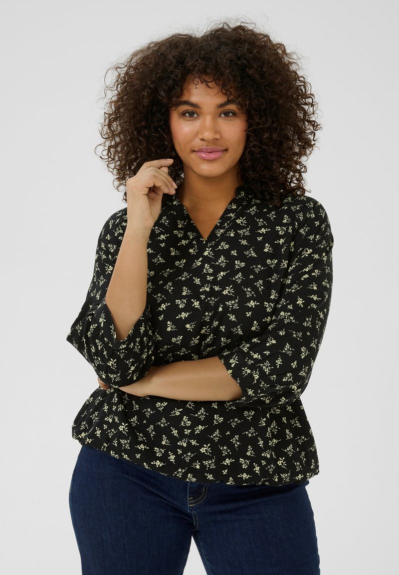 Blouse peplum noire avec un motif floral blanc, décolleté en V et manches 3/4. Le tissu a une texture douce, adaptée à un usage décontracté.