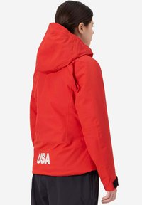 Giacca impermeabile rossa con cappuccio, dotata di maniche con cerniere e di un logo bianco "USA" sul retro, dalla texture liscia e dal design aderente.