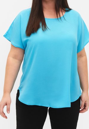 Femme portant un chemisier turquoise à manches courtes et amples et un pantalon noir, debout devant un fond clair uni.