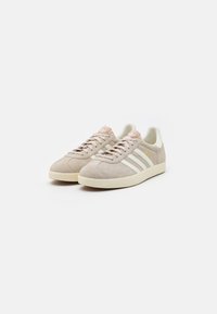 Beige suede sneakers with white three-stripe design, light pink accents on the tongue, and cream rubber sole. Classic low-top shape.