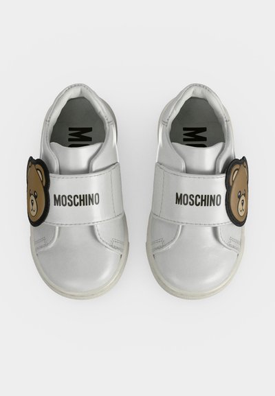Детски бели маратонки Moschino с велкро каишки и декорация на мече на обикновен фон.