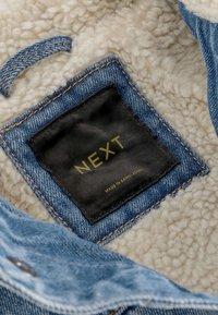 Džínová bunda s krémovou fleecovou podšívkou a čiernou prešitou nášivkou s nápisom "NEXT" a "Made in Bangladesh."