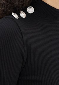 Czarny prążkowany top z dzianiny, odsłaniający ramiona, z trzema ozdobnymi okrągłymi srebrnymi guzikami w rzędzie przy kołnierzu.