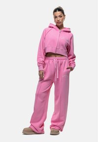 Roze cropped hoodie met rits aan de voorkant en logo, gecombineerd met losse roze broek met elastische taille en trekkoord. Zachte textuur.