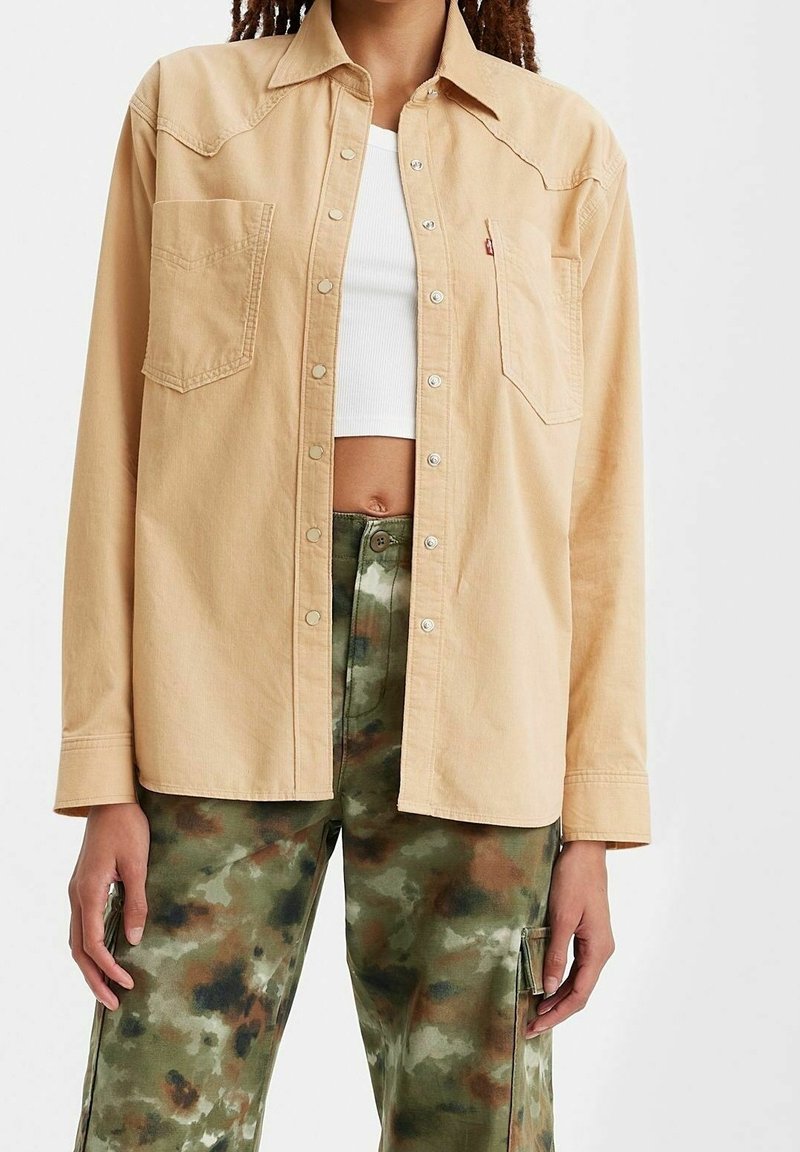 Personne portant une chemise beige boutonnée par-dessus un crop top blanc et un pantalon cargo camouflage vert.
