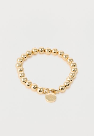 Bracelet en perles dorées avec fermoir à bascule rond et petit charme circulaire gravé avec "RL" sur un fond blanc.
