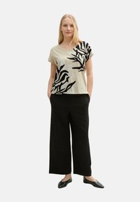 Beige top met V-hals en korte mouwen, versierd met een zwart organisch bladmotief. Gecombineerd met zwarte broek met brede pijpen en zwarte ballerina's, wat een casual uitstraling creëert.