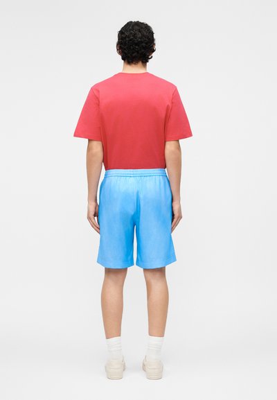 Άνδρας με σγουρά μαλλιά φοράει κόκκινο μπλουζάκι, ανοιχτό μπλε σορτς, λευκές κάλτσες και λευκά αθλητικά, στέκεται με την πλάτη στην κάμερα πάνω σε μονόχρωμο φόντο.