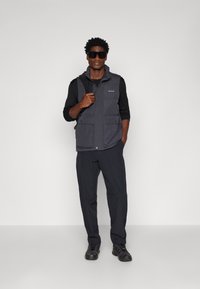 Dark gray sleeveless vest with two front pockets and a zipper, paired with dark long-sleeve shirt and pants, and black athletic shoes.