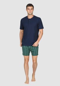 Maglietta henley a maniche corte blu navy con tre bottoni, abbinata a pantaloni corti verdi con un piccolo motivo di barche bianche. Modello scalzo in posizione eretta.