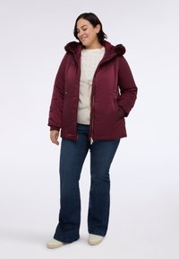 Burgundowa kurtka puchowa z odpinanym futrzanym kapturem, kieszeniami na zamek błyskawiczny i zamkiem z przodu. W zestawie z kremowym swetrem i dżinsami w stylu dzwonów.