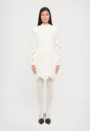 White lace dress with floral pattern, long puffed sleeves, high collar, and scalloped hem. Worn with nude tights and pointy black shoes.
