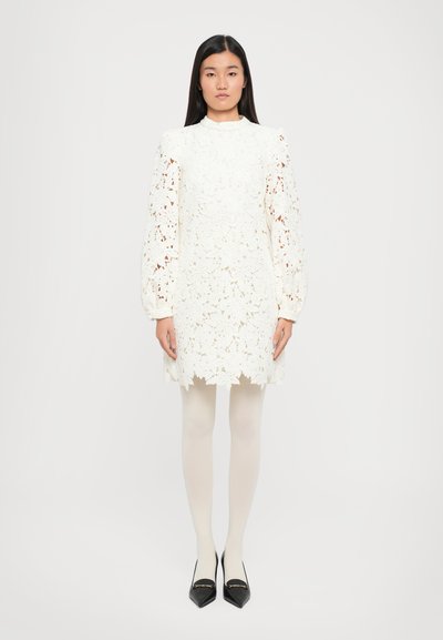 Robe en dentelle blanche avec un motif floral, manches longues bouffantes, col montant et ourlet festonné. Portée avec des collants couleur chair et des chaussures noires pointues.