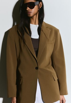 Žena odjevena u preveliki smeđi sako, bijelu majicu, tamnosmeđi top i velike crne sunčane naočale, na bijeloj pozadini sa sjenom.