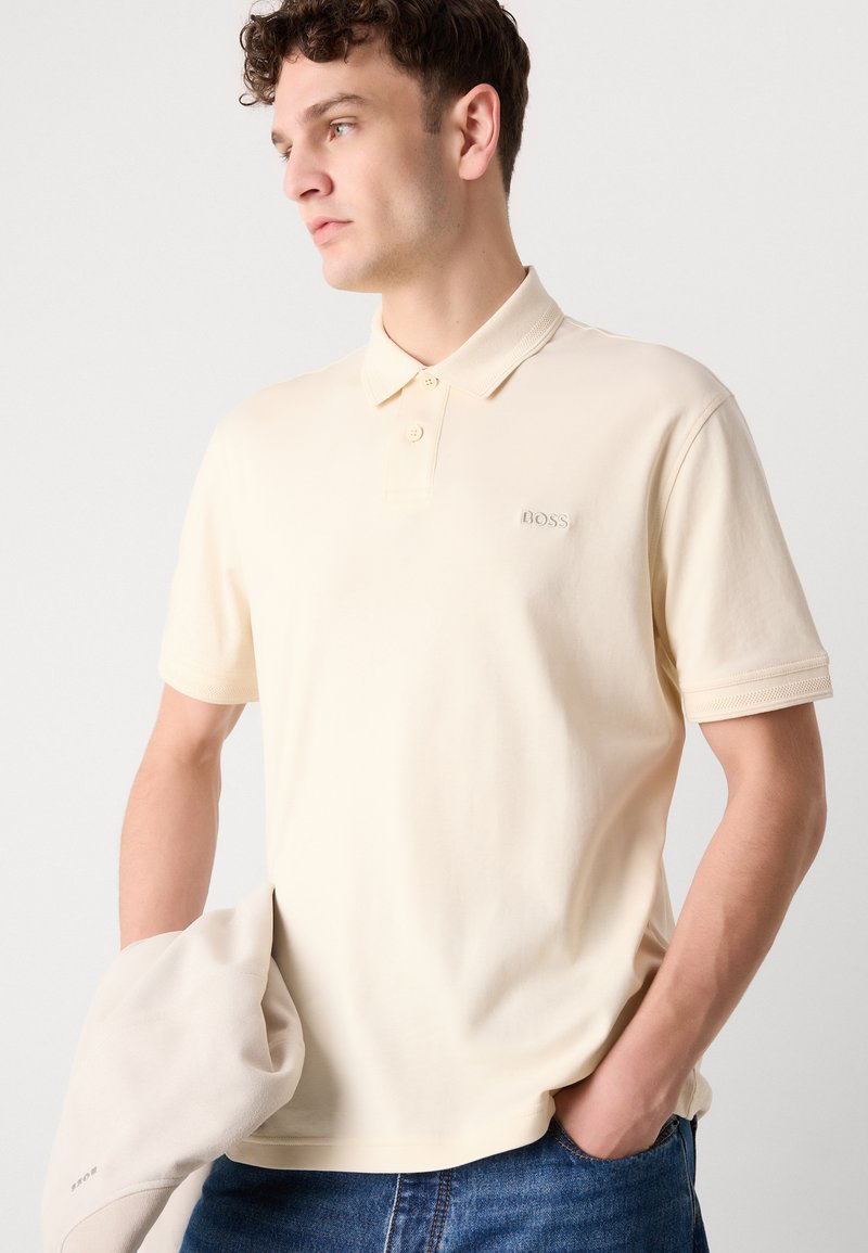 Polo crème avec col classique, manches courtes et logo ton sur ton brodé sur la poitrine. Texture en coton douce et coupe décontractée.