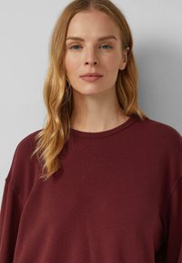 Donna bionda con capelli mossi indossa un maglione bordeaux, guardando dritto davanti a sé su uno sfondo grigio chiaro e uniforme.