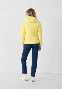 Giacca di piumino gialla con cappuccio, design trapuntato e forma aderente. Abbinata a jeans blu e sneakers bianche. Vista posteriore.