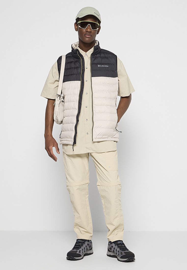Gilet imbottito leggero in nero e crema, camicia beige a maniche corte, pantaloni beige e scarpe da trekking grigie. Gli accessori includono occhiali da sole e un cappellino.