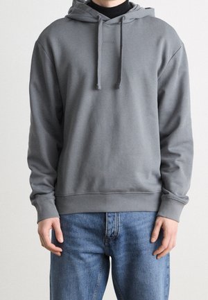 Person trägt einen grauen Kapuzenpullover mit Kordelzug und blaue Jeans, steht vor einem schlichten hellen Hintergrund.