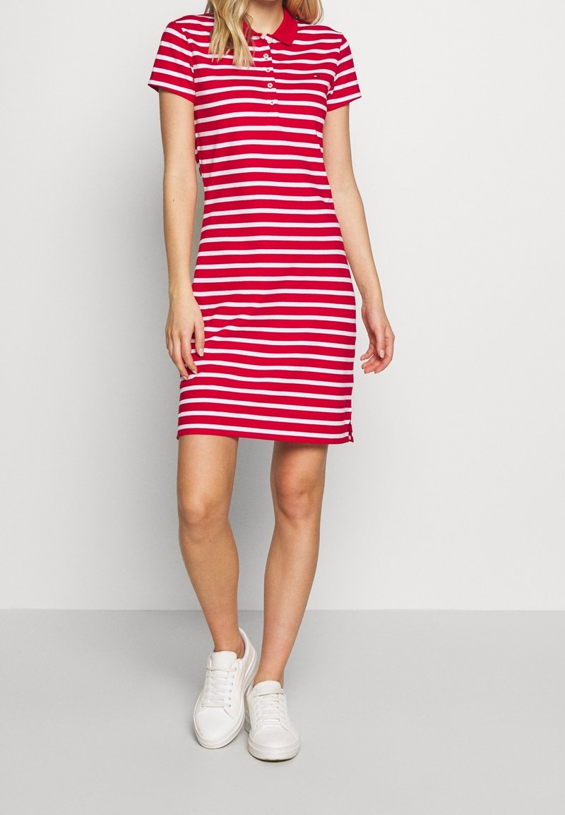 Femme portant une robe polo à rayures horizontales rouges et blanches et des baskets blanches, debout devant un fond uni clair.