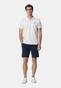 Polo bianco a maniche corte con un piccolo design ricamato sul lato sinistro del petto, abbinato a pantaloni corti navy e sneakers bianche.