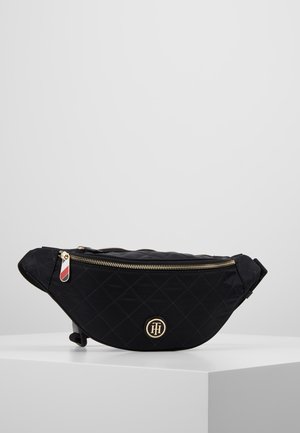 Zwarte gewatteerde stoffen heuptas met gouden rits en logo-embleem, tentoongesteld op een witte voetstuk tegen een grijze achtergrond.