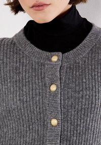 Frau, die eine graue gerippte Strickjacke mit drei strukturierten goldenen Knöpfen über einem schwarzen Rollkragenpullover trägt, von Kinn bis zur oberen Brust gezeigt.