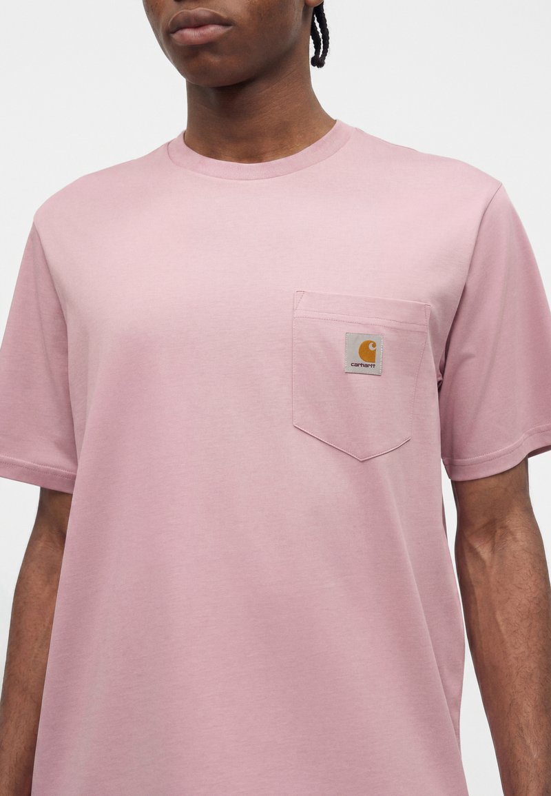 Person wearing a light pink short-sleeve t-shirt with a small Carhartt logo patch on the left chest pocket against a plain background.