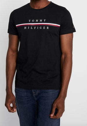 T-shirt en coton noir avec col rond, manches courtes et une bande horizontale blanche, rouge et bleue proéminente avec le nom de la marque.