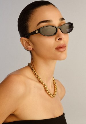 Mujer con el pelo peinado hacia atrás, lleva gafas de sol ovaladas negras, un collar grueso de cadena dorada y un top negro sin tirantes, sobre un fondo neutro.