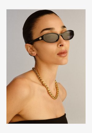 Frau mit zurückgestrichenen Haaren, die schwarze ovale Sonnenbrillen, eine dicke goldene Halskette und ein schwarzes trägerloses Oberteil vor neutralem Hintergrund trägt.