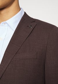 Blazer en lin marron avec une finition texturée et un pin's logo noir sur le revers. Porté sur une chemise à col bleu clair.