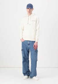 Krem hettegenser med snøring og frontlomme, sammen med lyseblå baggy cargo jeans med sidelommer, og hvite joggesko. Blå caps.