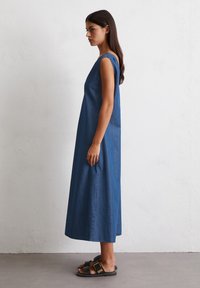 Ermeløs, knelang blå denimkjole med en avslappet passform, featuring V-hals og jevn tekstur. Kombinert med flate svarte sandaler.