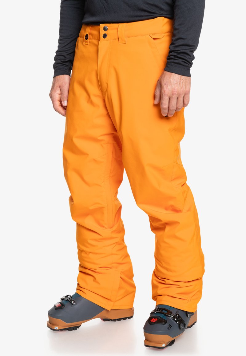 Jasno-pomarańczowe spodnie narciarskie o luźnym kroju, z dwoma przednimi guzikami i gładką, wodoodporną fakturą. Noszone na szarych butach narciarskich.