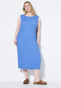Vestido midi sin mangas de color azul con tejido texturizado que presenta un patrón ondulado. Combinado con zapatos marrones tipo slip-on con acento de cadena.