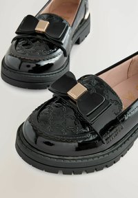 Schwarze Lackleder-Lederhosen mit einem strukturierten Design, ausgestattet mit einer auffälligen Schleifenakzent und goldenen Beschlägen an der Vorderseite. Gummisohle.