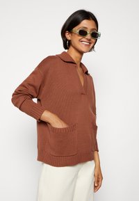 Brun strikket sweater med V-udskæring, krave og to frontlommer. Ribbet manchetter og kantbånd, struktureret materiale. Lyse bukser.